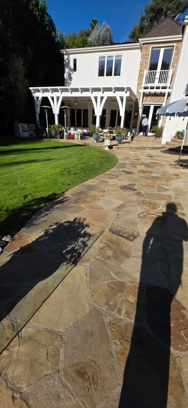 Daytime view of a flagstone patio and green lawn curving toward a white pergola attached to the house