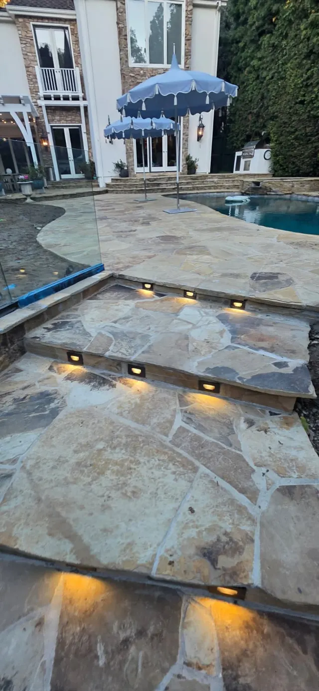 Pool edge and flagstone patio with uplit stone steps and blue patio umbrellas in front of the house at dusk