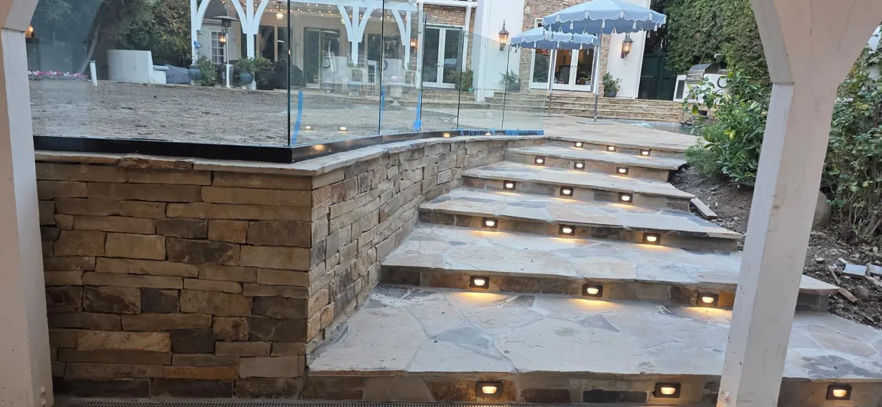 Stacked stone retaining wall with a glass railing above uplit flagstone stairs, leading up to a patio with a pergola and the house beyond